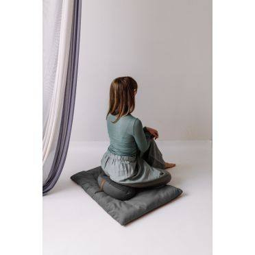 Kobieta w zielonej bluzce i szarej spódnicy siedzi tyłem na szarej poduszce do medytacji w kształcie fasolki, na dopasowanej macie. W tle zwiewna tkanina i biała ściana.